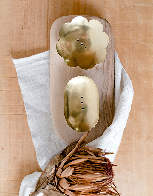 Nos deux modèles de porte savon doré en laiton, posés sur une planche en noyer avec fleur séchée à côté